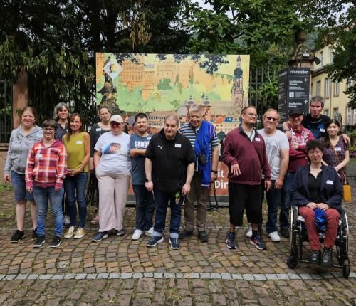 Interessierte Podcast-Teilnehmerinnen und Teilnehmer der Kunstgruppe der Lebenshilfe Heidelberg e.V. und des Heidelberger Beirats von Menschen mit Behinderung. Willkommen im Museumshof: interessierte Podcast-Teilnehmerinnen und Teilnehmer der Kunstgruppe der Lebenshilfe Heidelberg e.V. und des Heidelberger Beirats von Menschen mit Behinderung. stehen vor einer Werbetafel des Museums.